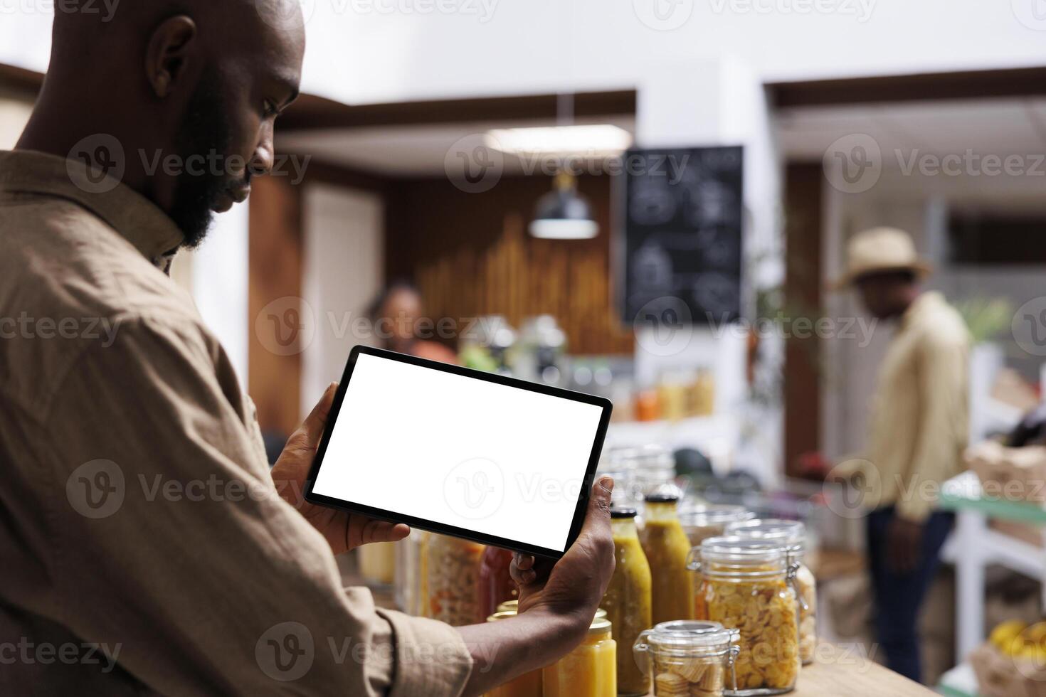 An African American guy consumer holds a phone tablet horizontally with an isolated chromakey mockup template. On wooden shelves, the store sells fresh, eco friendly products. photo