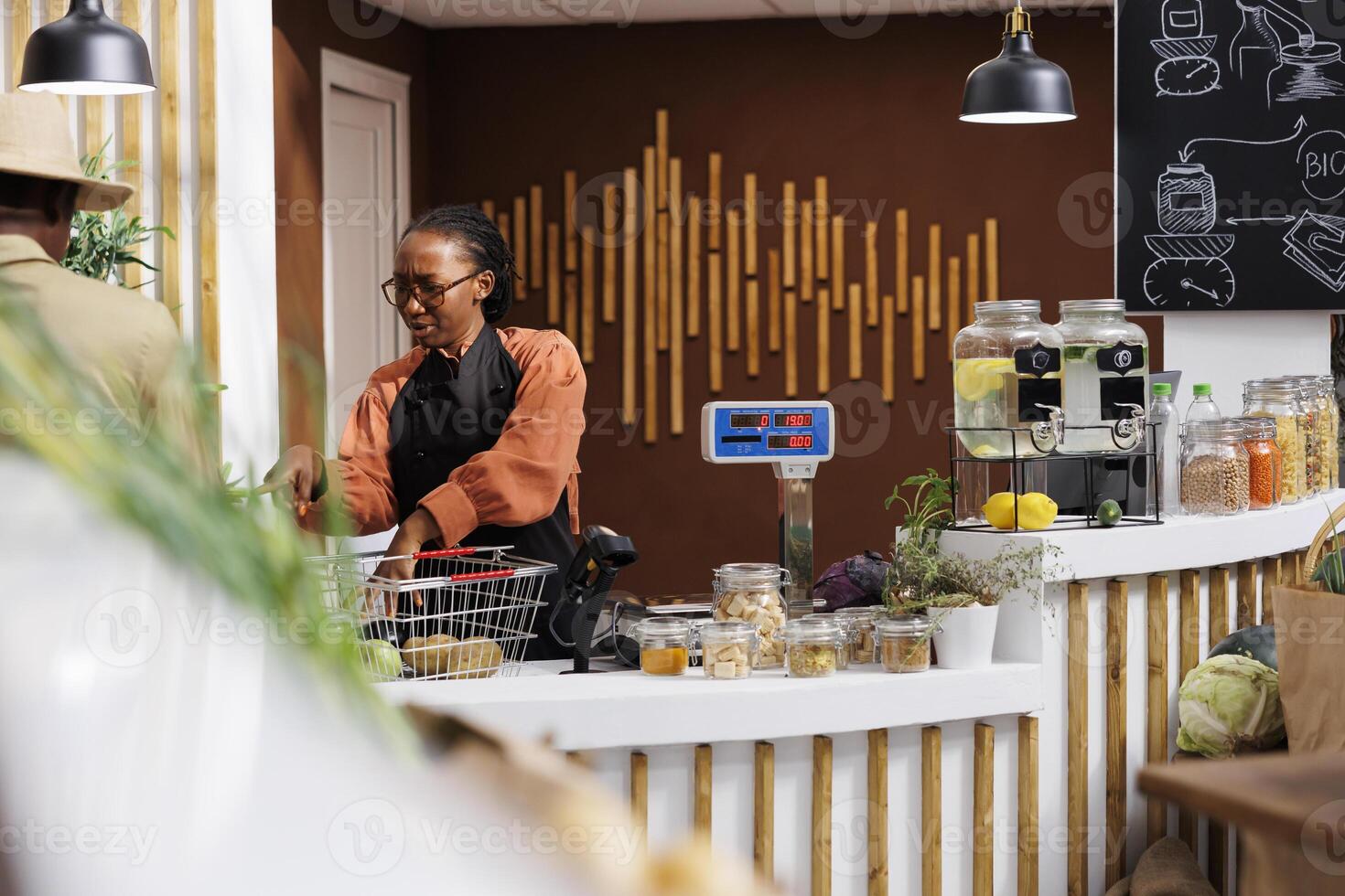 African American female cashier in a black apron gesturing to something and assisting the male customer. Young black woman at payment counter removing items from basket for weighing. photo