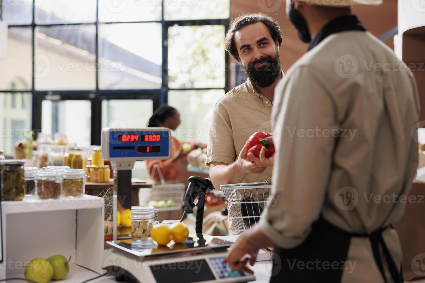 Man with an apron checking the weight of produce using modern scale and selling fresh items to caucasian customer. Local grocery store vendor weighing fruits and vegetables for client. photo