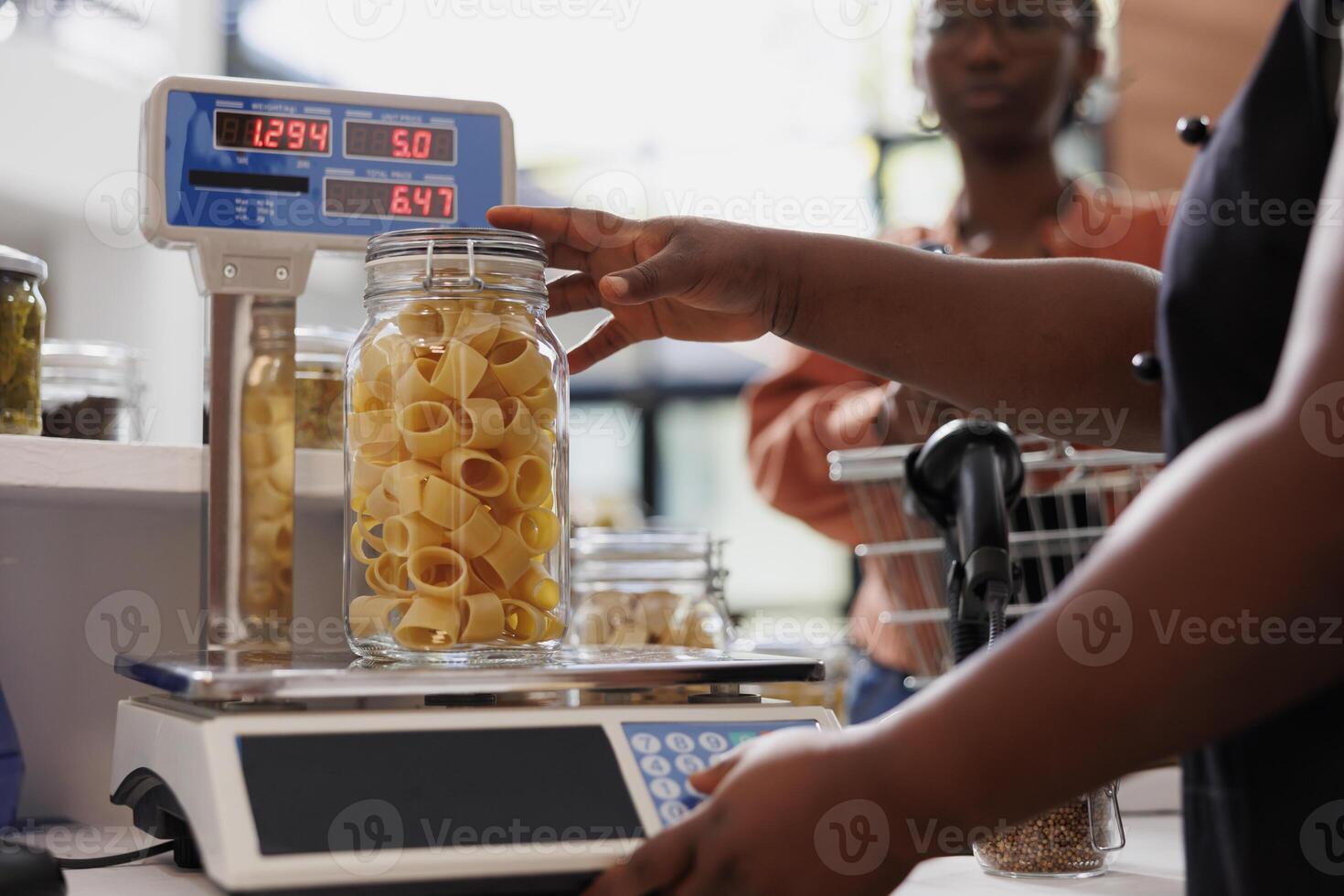 Closeup of african american vendor using digital scale to measure weight of fresh organic product in glass container. Detailed view of merchant weighing jar of bio pasta while client waits at counter. photo