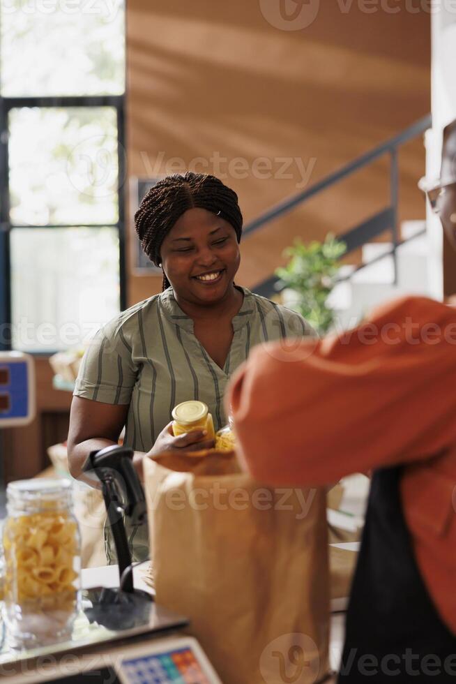 Cheerful female customer with locally grown produce at the checkout counter of grocery store. Vendor puts freshly harvested fruits and vegetables in a paper bag as african american consumer waits. photo