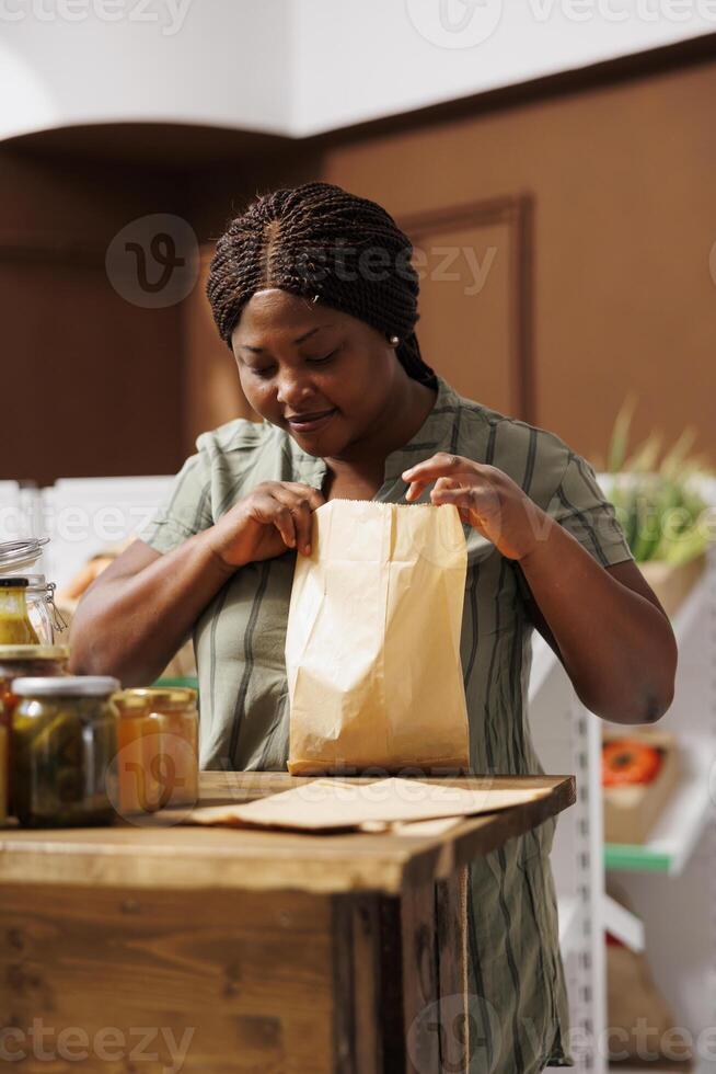 Black woman searching for fresh and healthy produce in store filled with eco-friendly containers and jars, promoting sustainable and zero waste living. Female client checking inside paper bag. photo