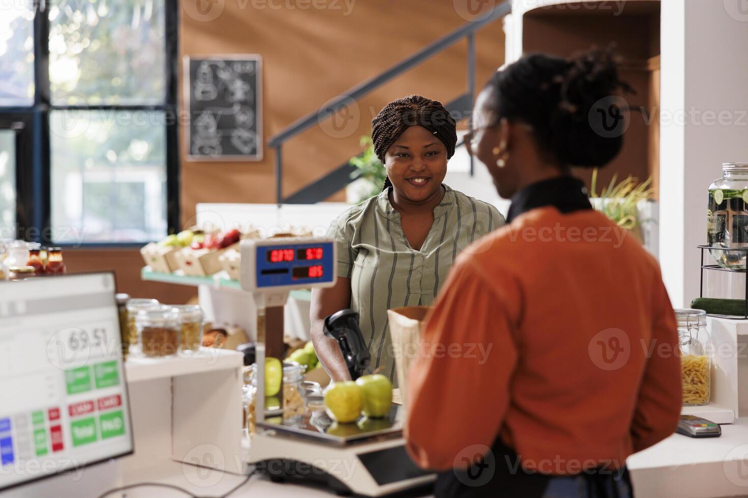 Smiling black lady receiving assistance from African American vendor at the checkout counter with her locally grown produce. Customer at cashier desk in zero waster store purchasing organic products. photo