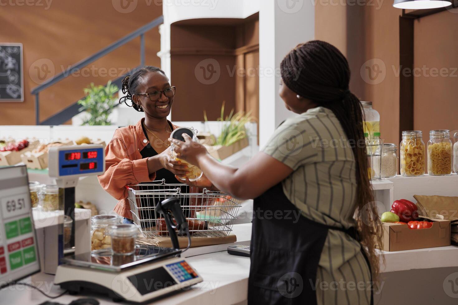 African American vendor standing behind counter serving female customer, offering locally grown produce for healthy lifestyle. Young consumer handing over glass jar of pasta to seller for weighing. photo