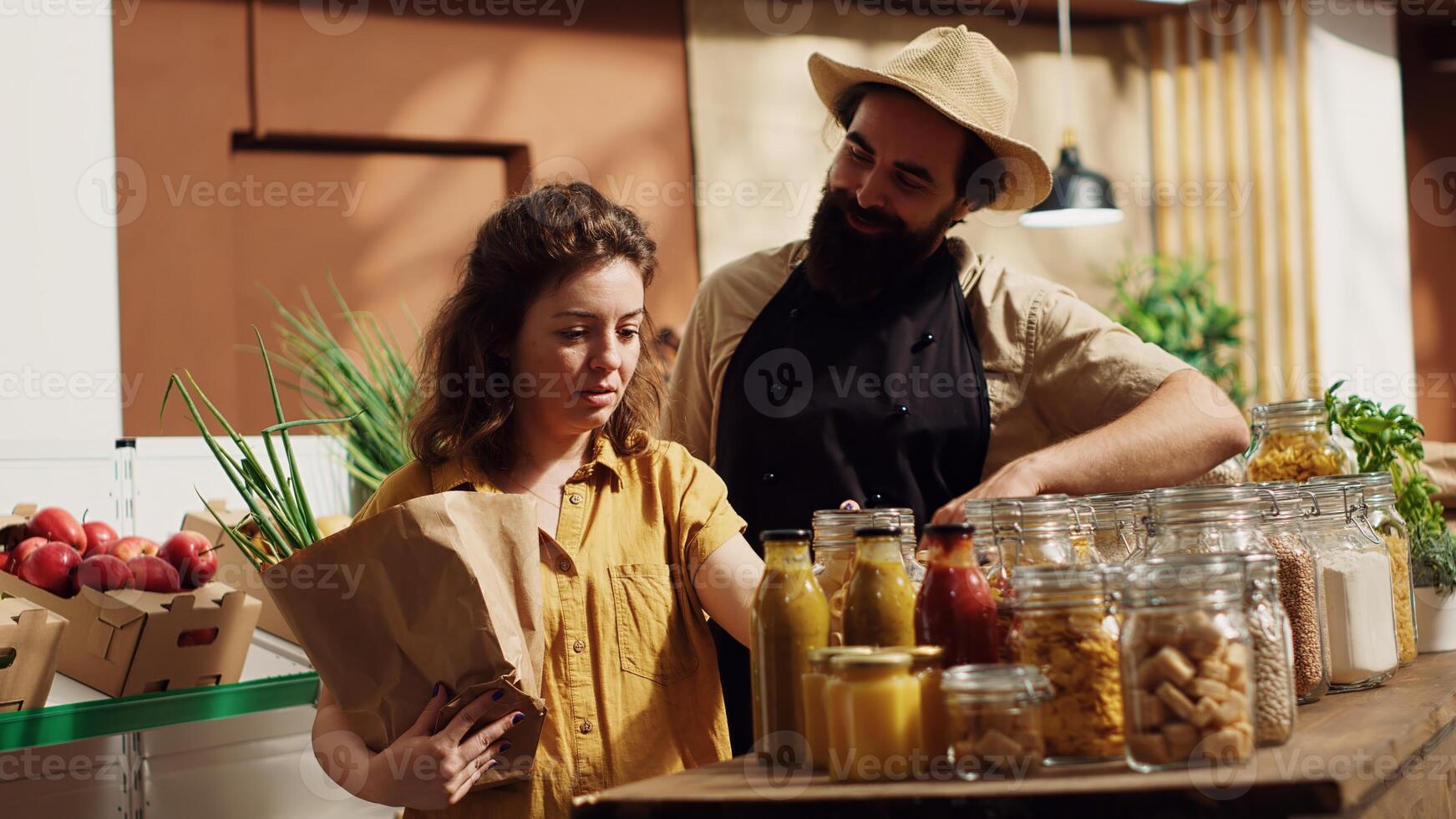 Woman adding pesticides free pantry essentials and vegetables in paper bag, getting product recommendations from vendor in zero waste supermarket. Trader helping client in bio local grocery shop photo