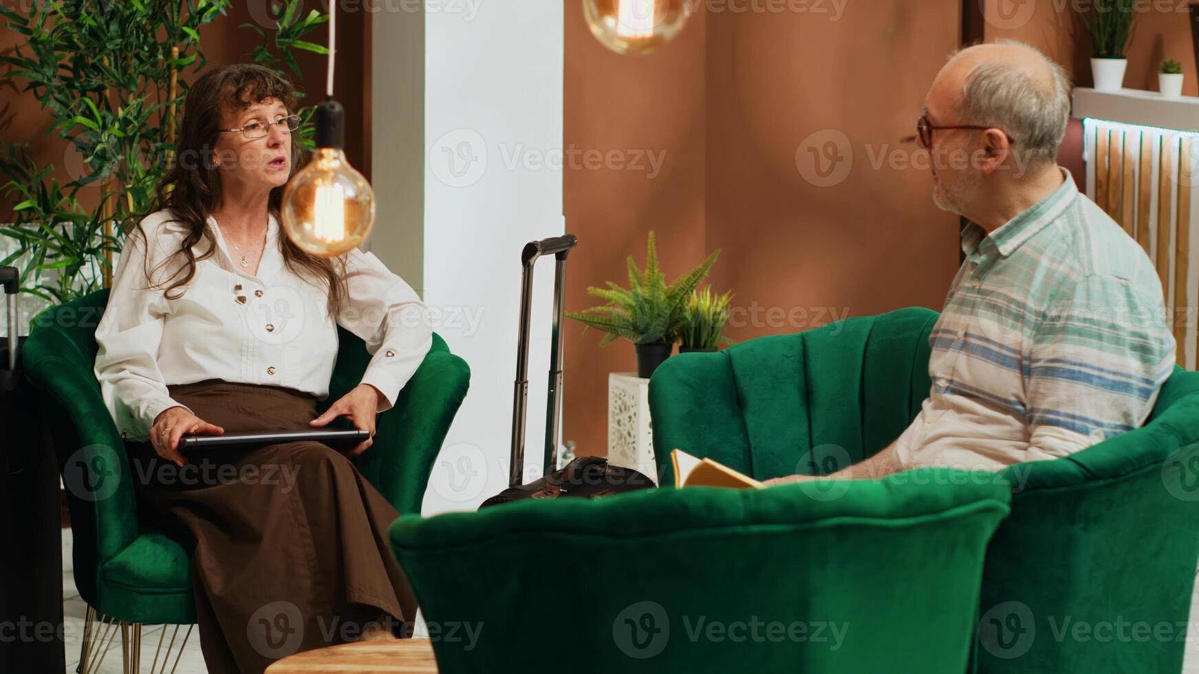 People passing time in hotel lobby, travelling on retirement holiday abroad and waiting for check in in lounge area. Older woman looks at famous visiting places on laptop and man enjoying book. photo