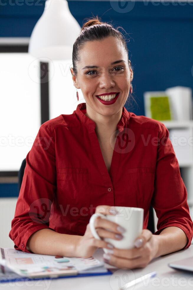 Portrait of cheerful happy excited business leadership smiling looking at camera. Entrepreneur with financial charts on clipboard on office desk in corporate workplace. Holding cup of coffee. photo