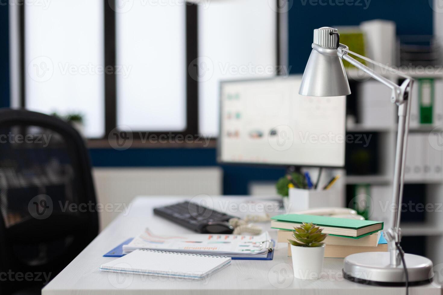 Closeup of lamp on white desk in empty modern business office. Notebook and clipboard with papers on table in startup workspace. Computer screen in indoors modern commercial workplace. photo