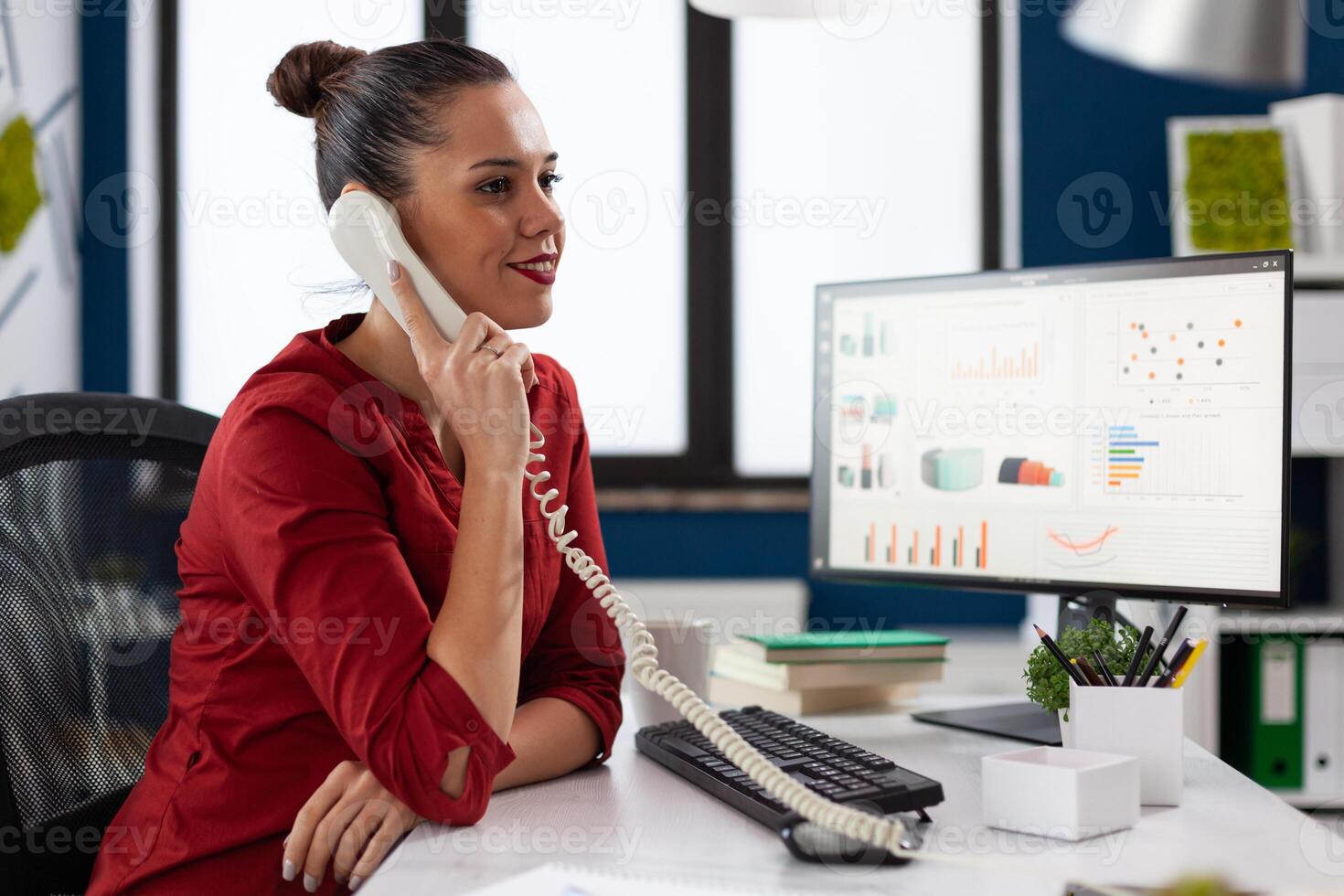 Smiling businesswoman in phonecall sitting at startup desk. Entrepreneur in red shirt listening to employee at the phone. Manager in office sitting in front of computer with charts. photo