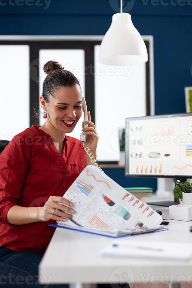 Smiling entrepreneur talking on the telephone explaining charts. Successful manager happy with business results in startup office. Employee in red shirt working at desk with clipboard and computer. photo