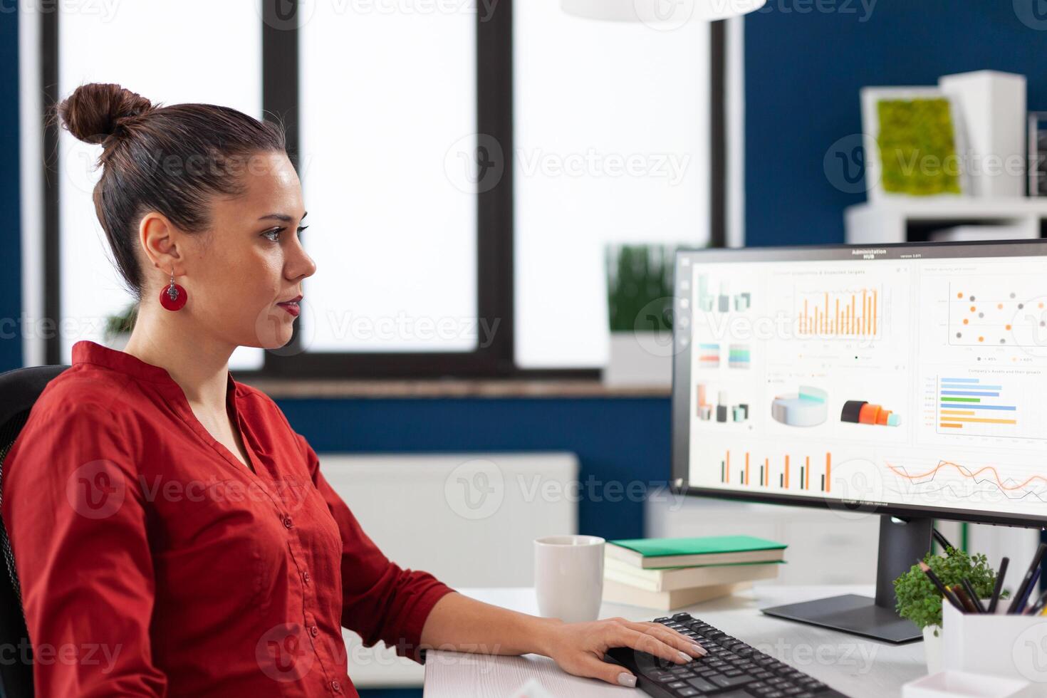 Confident entrepreneur looking at business results on computer screen. Businesswoman working at desk in startup office. Manager in red shirt typing on pc keyboard and looking at charts on display, photo
