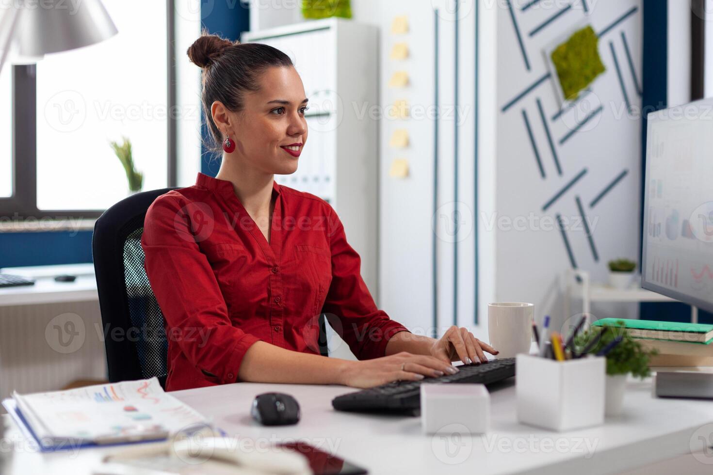 Smiling startup owner looking at computer monitor typing on keyboard. Small business manager in red shirt looking at charts on pc display. Employee sitting and working at desk in start up office. photo