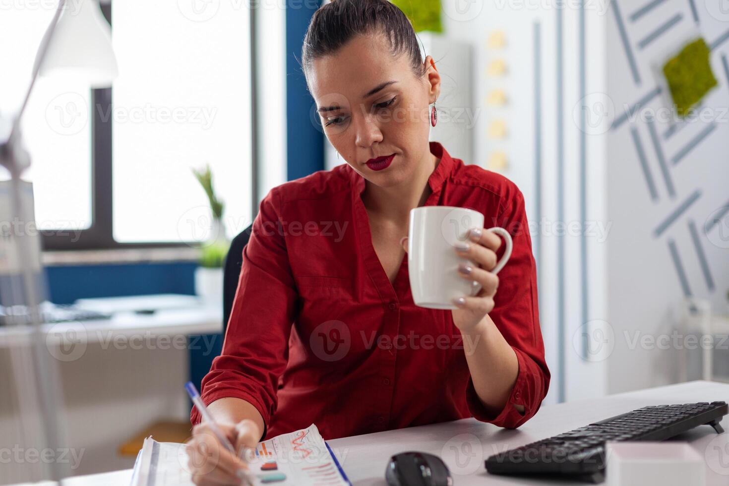 Startup manager holding cup writing on clipboard with chart. Entrepreneur in red shirt with pen in one hand working at desk in startup office. Focused employee analyzing small business data. photo