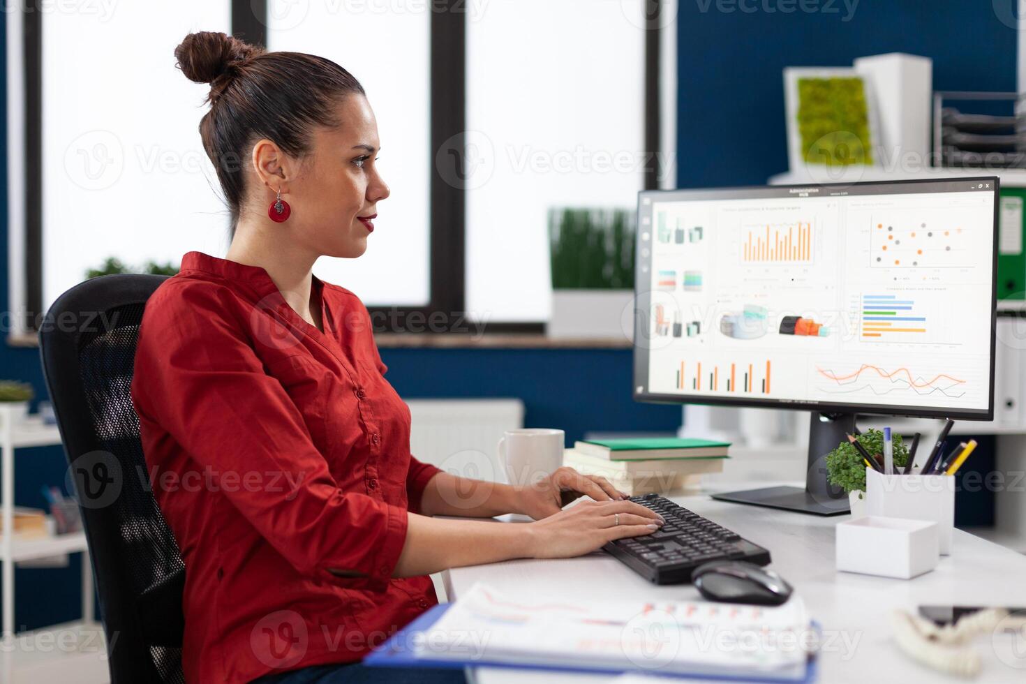 Startup manager in red shirt looking at charts on computer monitor. Smiling small business owner typing on keyboard at desk in start up office. Employee with clipboard analyzing graph and data. photo