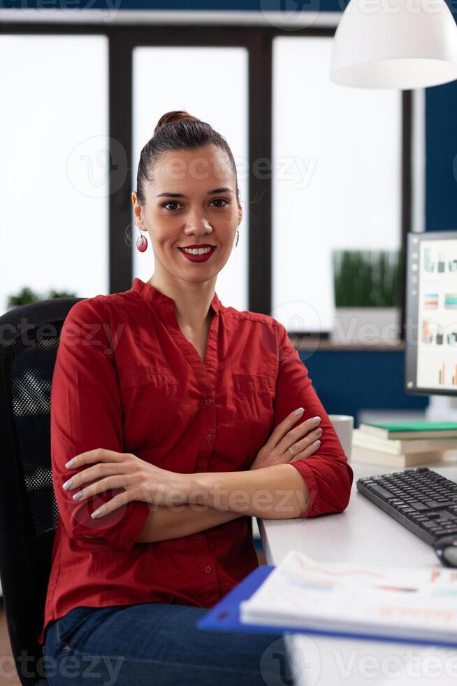 Portrait of entrepreneur sitting at desk in startup office. Businesswoman in red shirt looking confident in business workspace. Successful employee posing with crossed arms in front of desktop computer. photo