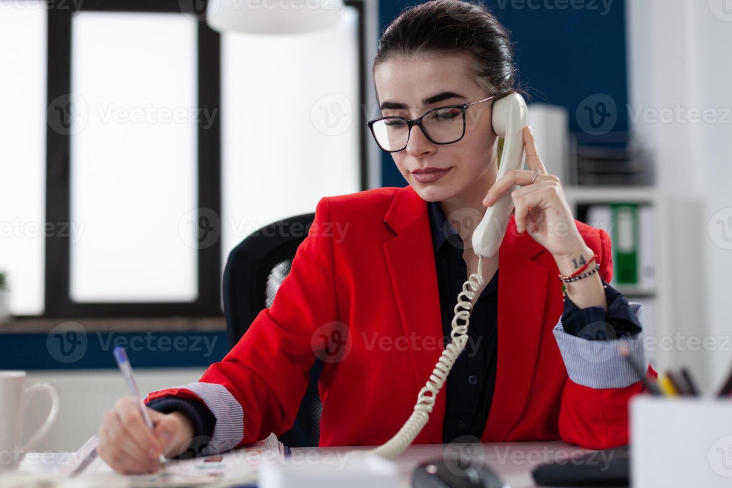 Entrepreneur taking notes in phonecall sitting at desk. Businesswoman with glasses working in startup office. Employee in red jacket writing on paper in telephone conversation. photo
