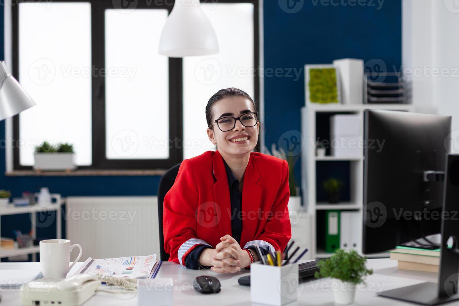 Portrait of businesswoman with glasses in startup office. Professional entrepreneur in red jacket looking confident in business workspace. Confident employee posing in front of desktop computer. photo