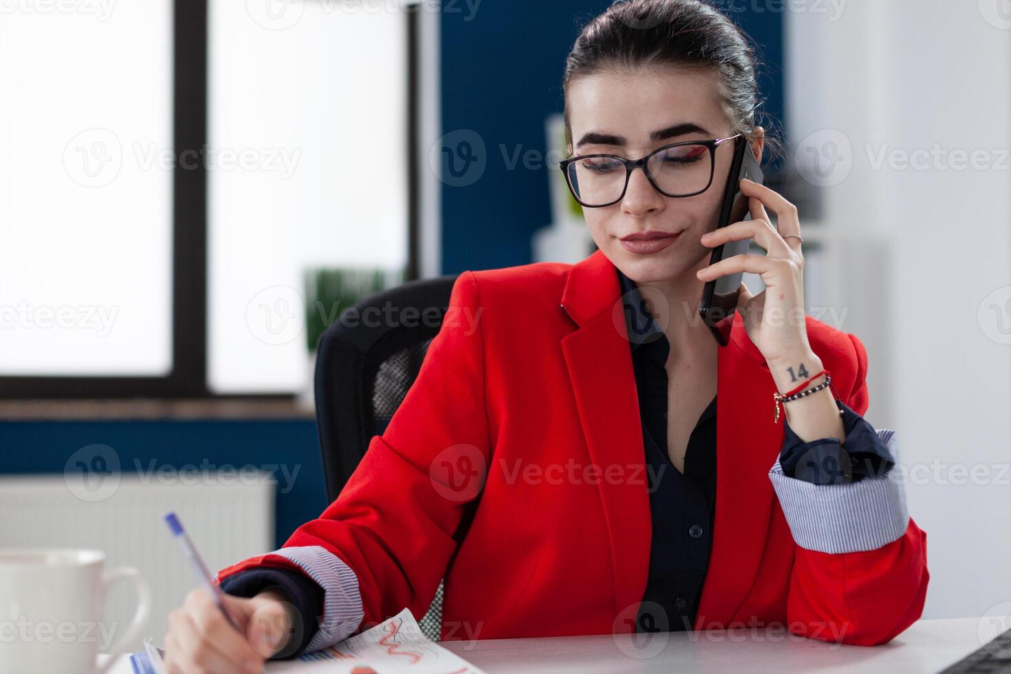Confident employee with glasses taking notes in phonecall sitting at desk. Small business owner in red jacket writing on paper in startup office. Entrepreneur taking notes in smartphone conversation. photo