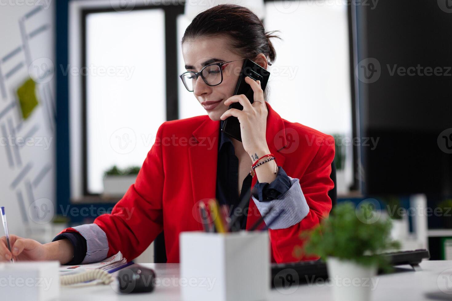 Businesswoman taking notes in smartphone phonecall sitting at desk. Entrepreneur with glasses working in startup office. Small business owner in red jacket writing on paper in startup enviroment. photo