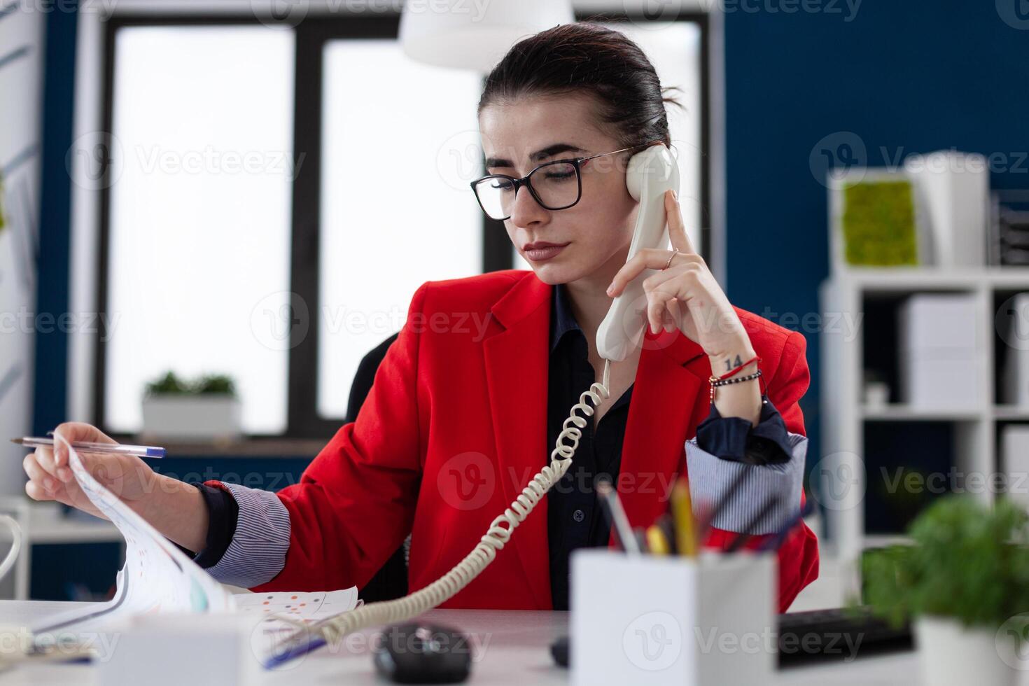 Businesswoman with glasses looking at papers in phonecall. Entrepreneur in red jacket sitting at desk analyzing clipboard with chart. Small business owner in startup office in phone conversation. photo