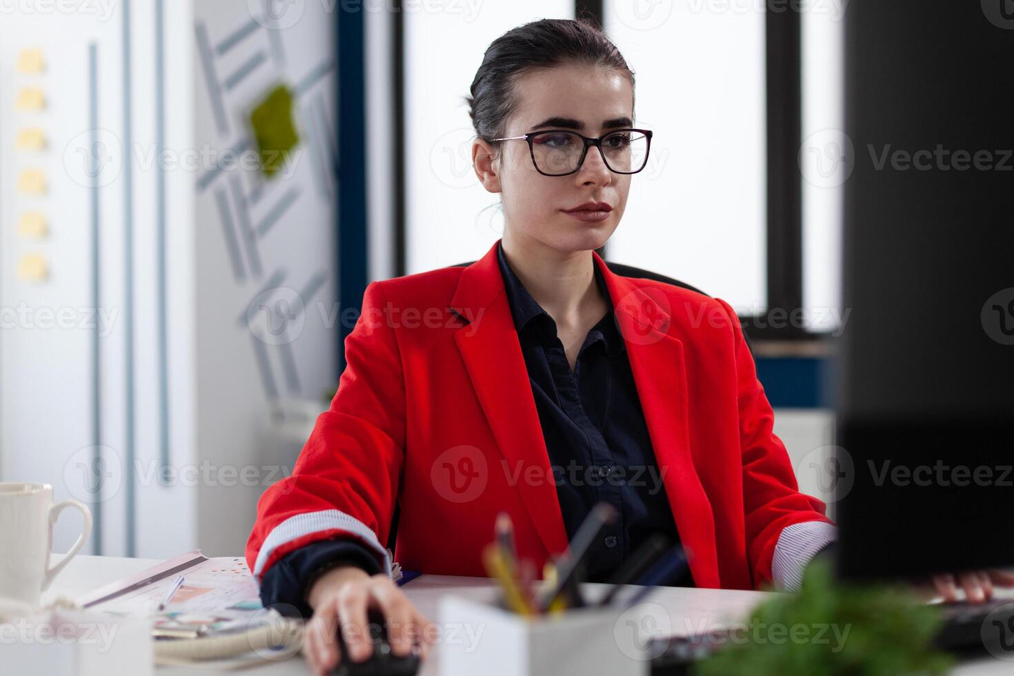 Confident small business owner with glasses working at desk in startup office. Businesswoman at workplace using desktop pc. Employee in red jacket scrolling content using computer mouse. photo