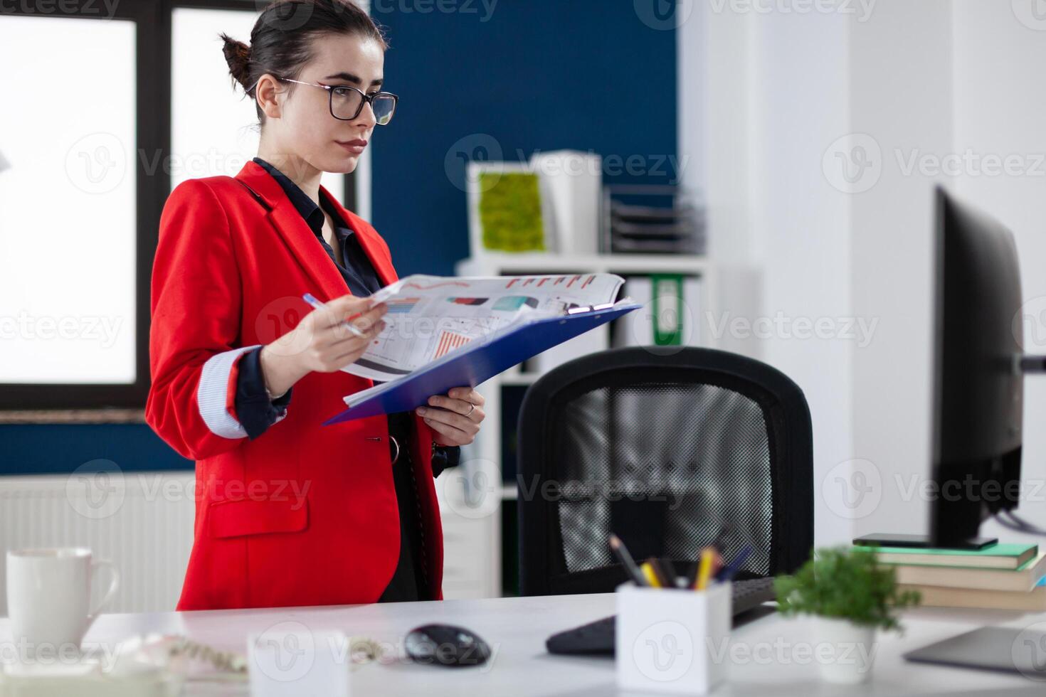 Businesswoman standing up next to desk holding clipboard with chart. Focused entrepreneur with glasses and red jacket in startup office looking at business results on computer screen. photo