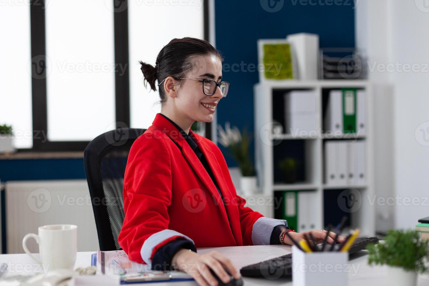 Smiling startup owner with glasses working at desk in office. Happy small business owner with glasses using dektop pc. Content entrepreneur in red jacket looking at computer screen. photo