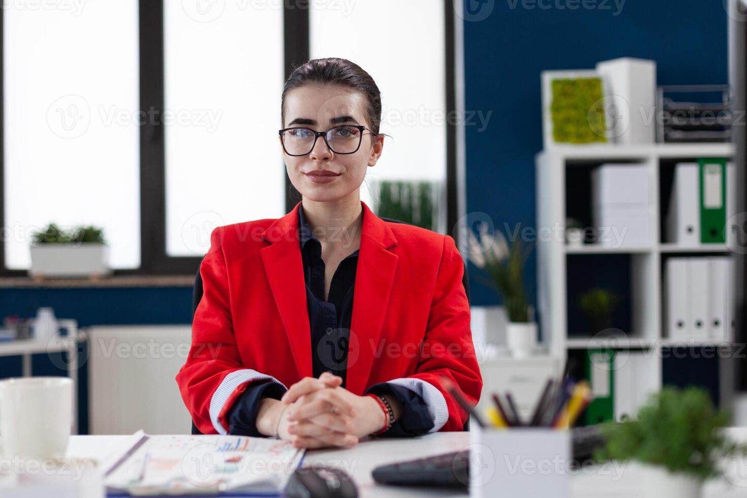 Portrait of entrepreneur with glasses in startup office. Professional businesswoman in red jacket looking confident in business workspace. Confident employee posing sitting at desk. photo