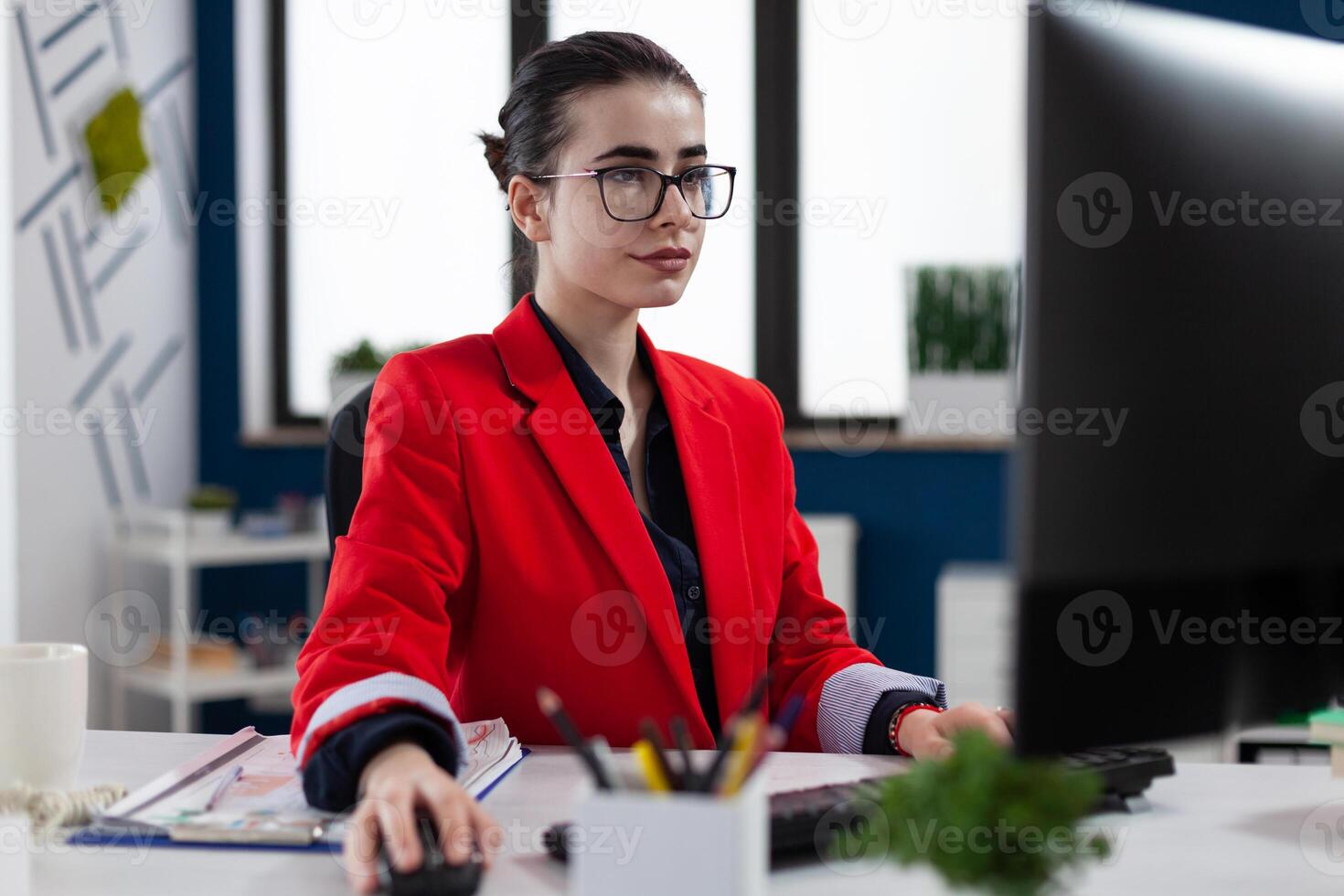 Small business owner with glasses working at desk in startup office. Businesswoman with glasses working in startup worpklace. Focused entrepreneur in red jacket looking at computer screen. photo