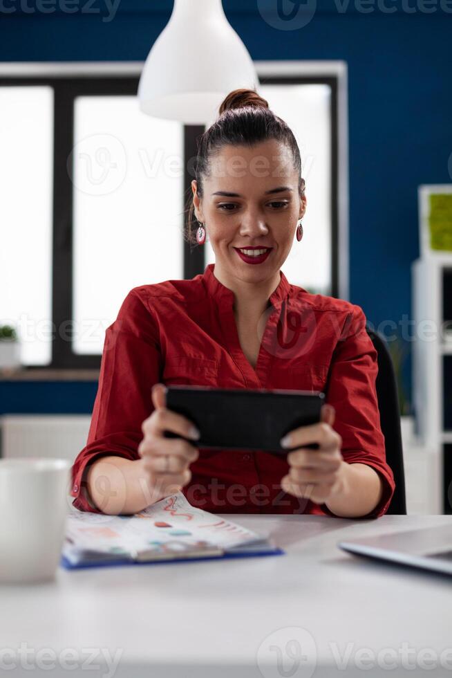 Businesswoman in red shirt sitting at desk holding smartphone. Employee with gadget streaming content on social media. Entrepreneur having a break playing mobile games at the startup office. photo