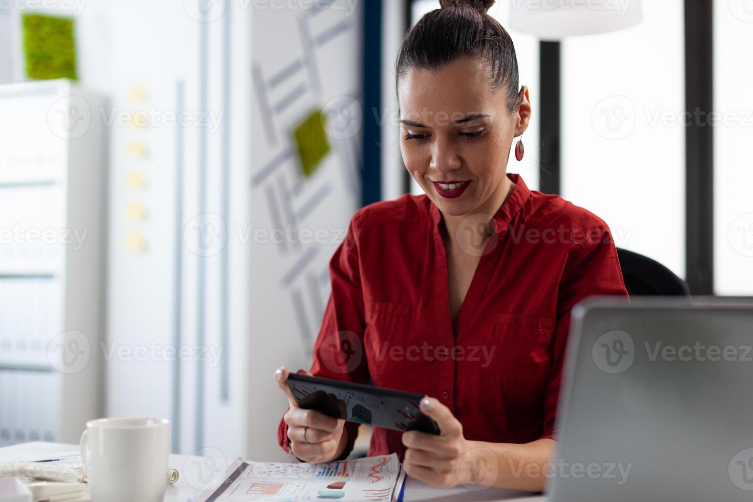 Smiling entrepreneur having a break playing mobile games at the startup office. Employee in red shirt sitting at desk holding smartphone. Businesswoman with gadget streaming content on social media. photo