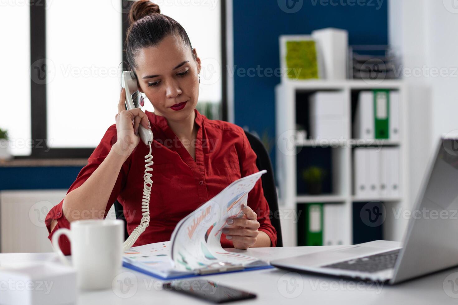 Entrepreneur in red shirt analyzing chart in phonecall. Businesswoman sitting at desk looking at clipboard with business papers. Employee in startup office in phone conversation. photo