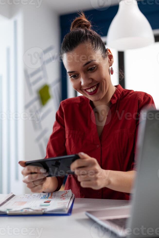 Employee in red shirt holding smartphone and looking at screen. Entrepreneur at the startup office in videocall conference. Businesswoman having a break playing casual mobile games online. photo