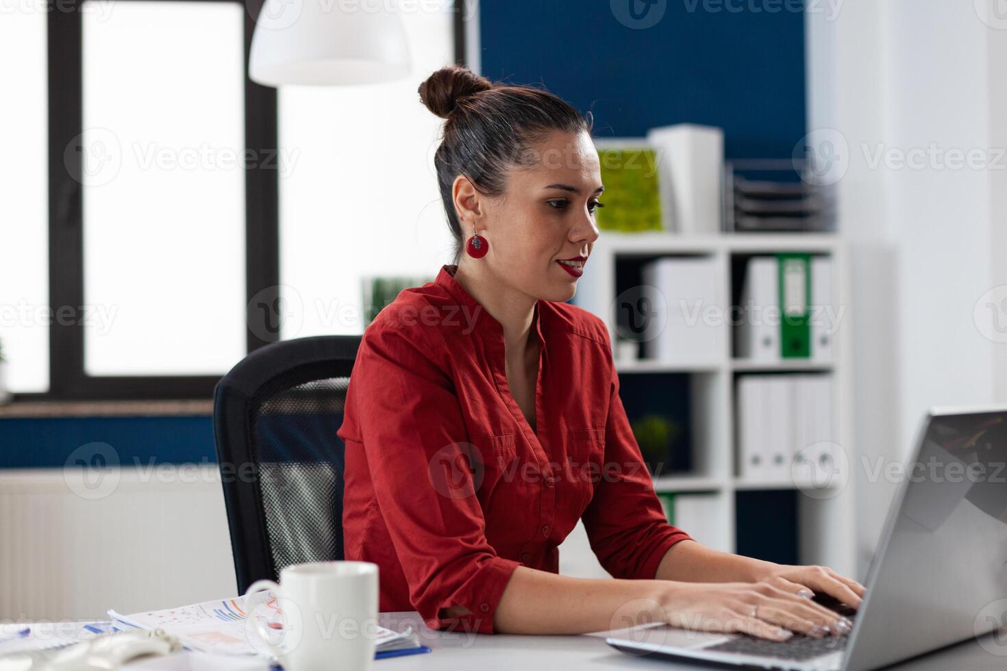 Confident businesswoman in red shirt working at desk in startup office. Business manager typing on laptop keyboard. Focused start up owner at workplace using notebook computer. photo