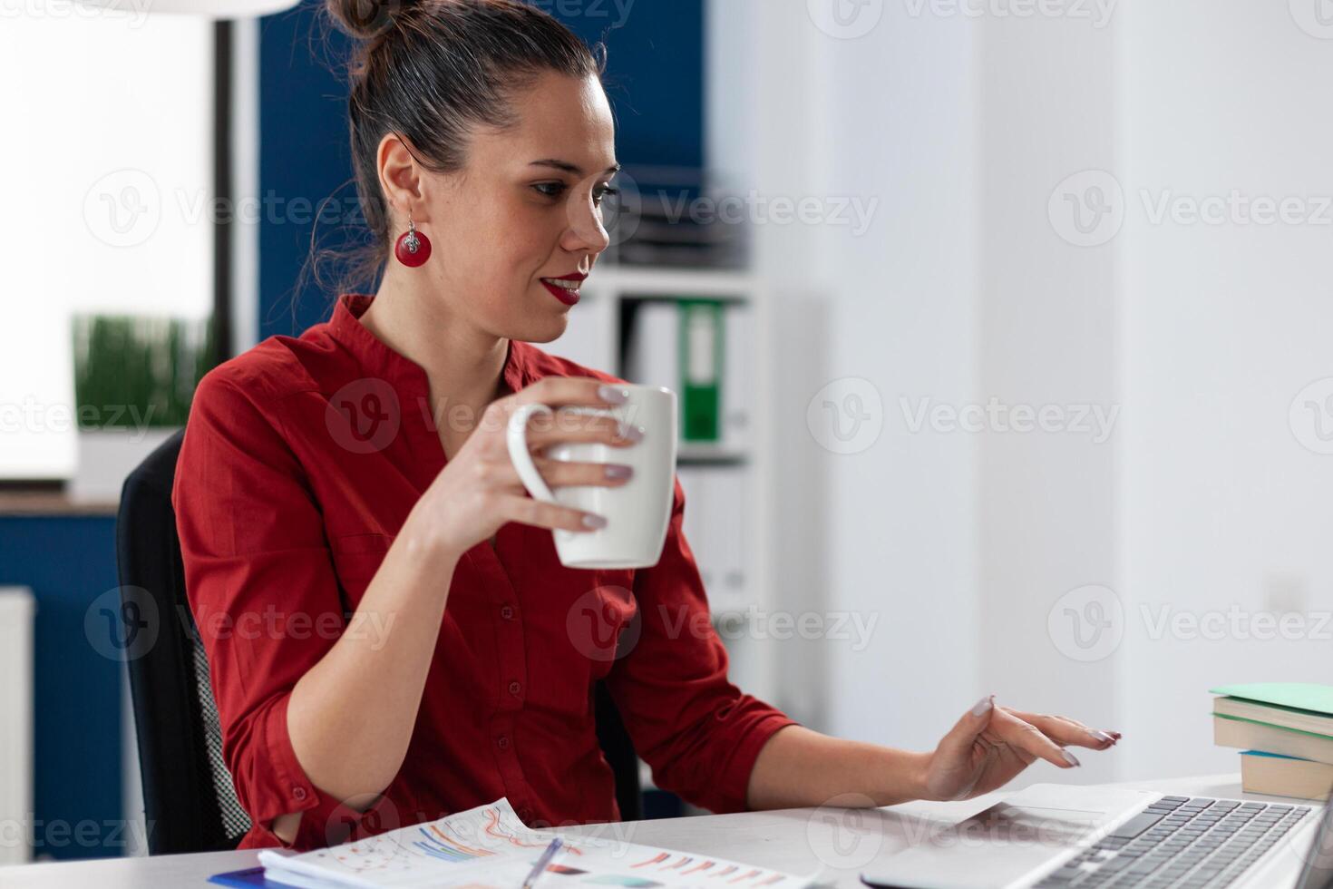 Startup manager working casually on laptop while holding white cup. Businesswoman in red shirt taking a coffee or tea break. Employee using notebook computer trackpad to browse internet. photo