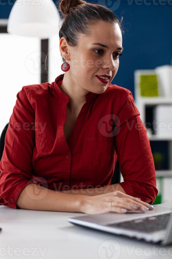 Smiling businesswoman in red shirt looking at laptop screen. Confident entrepreneur in call conference holding hand on touchpad. Small business owner watching content on social media. photo