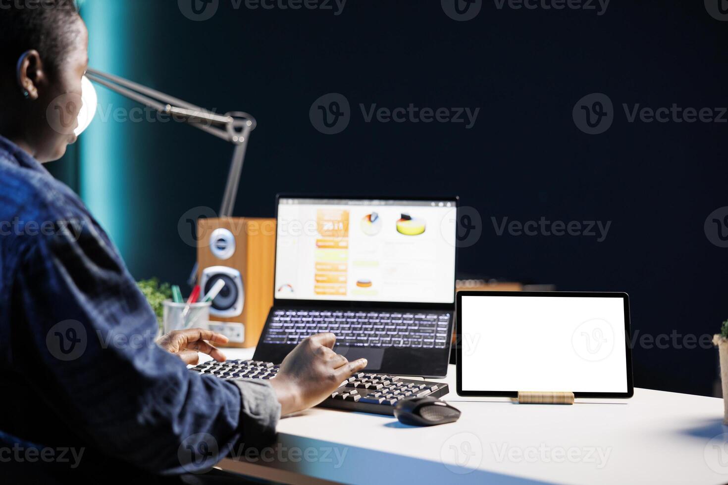 Black woman sitting at desk, tablet showing isolated white screen while she browses on her laptop. African american freelancer using her personal computer as a digital device displays mockup template. photo