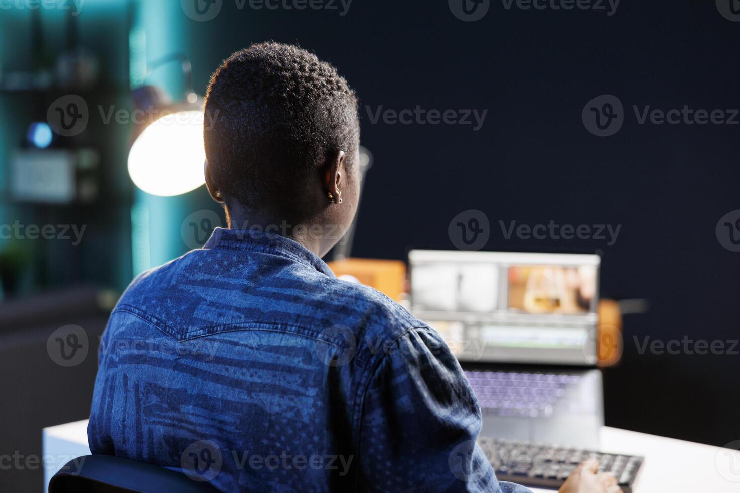 An African American woman edits footage on her laptop at her home office desk, immersed in creative work. Modern studio setup with digital device and software for productive filmmaking. photo