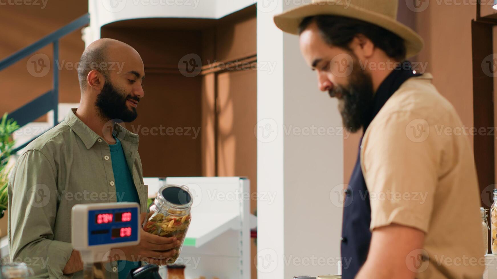 Man in apron checking weight of products using modern supermarket scale and sell fresh food to customer. Local grocery store owner weighting potatoes and pasta in reusable jar. Handheld shot. photo