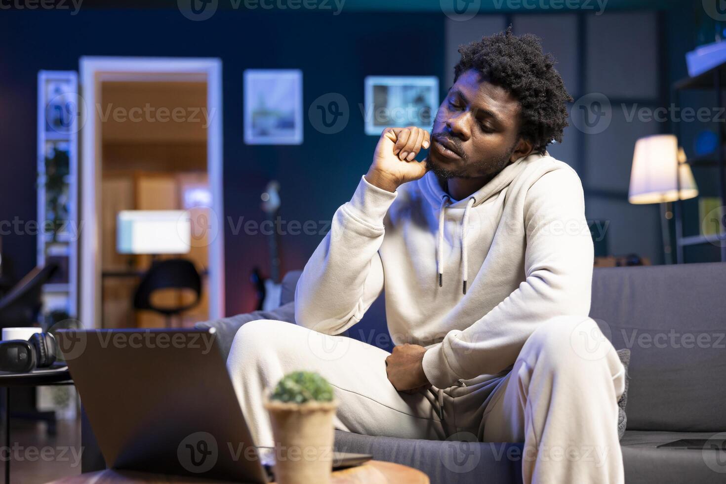 BIPOC bored man passing time at home by watching TV show on laptop. Tired person feeling apathetic in neon lit apartment at night while consuming uninteresting media content photo
