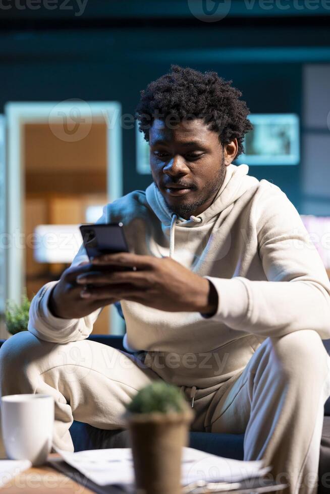 Man texting internet friends using mobile phone in warm stylish home, having fun. Joyful person relaxing in apartment by scrolling on smartphone, entranced by social media activity photo