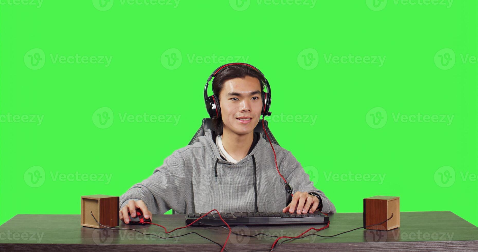 POV of asian guy playing at pc desktop, sitting against full body greenscreen backdrop. Young gamer having fun with players online in gaming competition, wearing headphones at workstation. photo