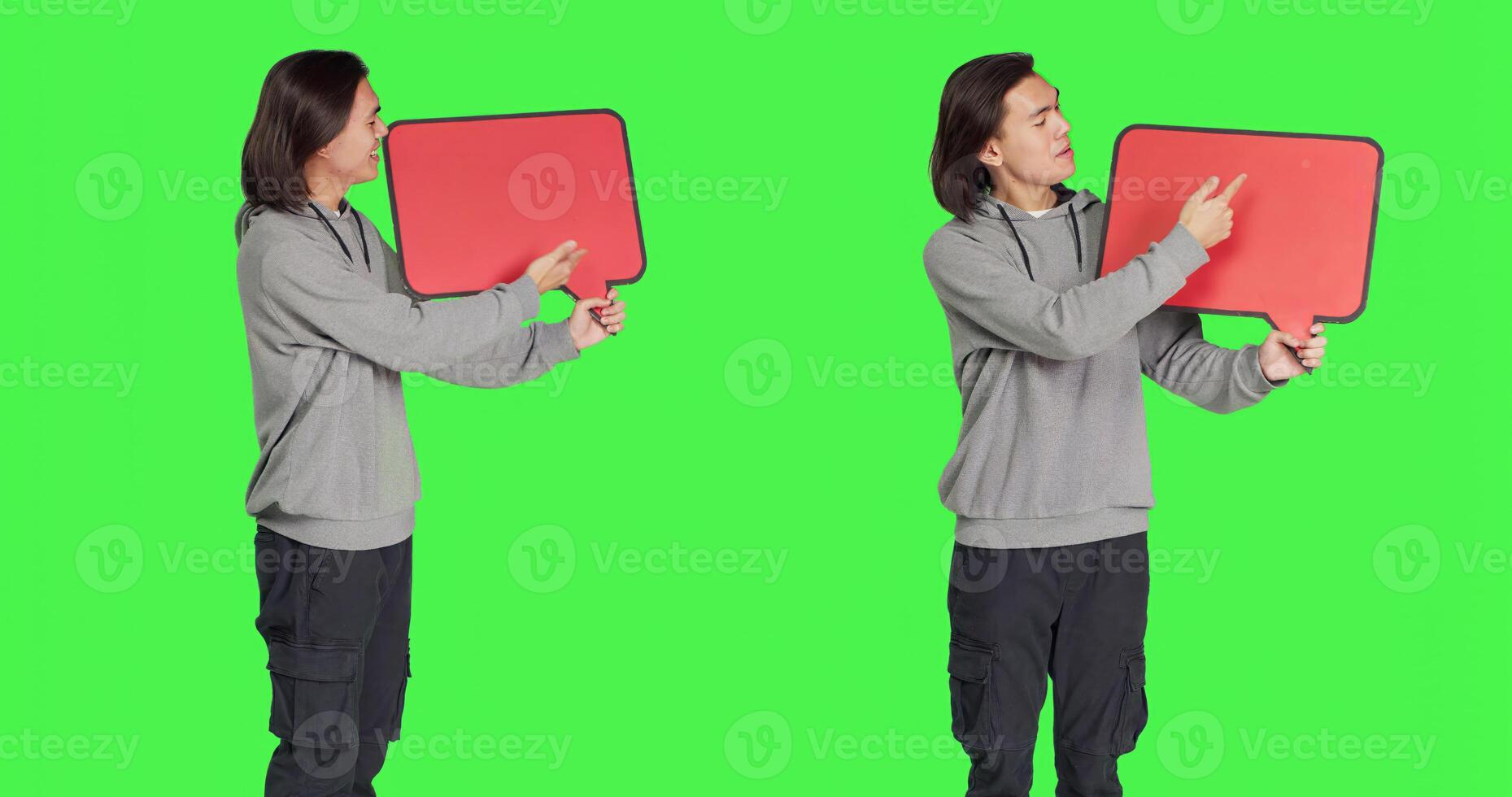 Young adult poses with speech bubble, presenting empty billboard with copyspace against greenscreen backdrop. Asian male model holding cardboard for marketing advertisement in studio. photo