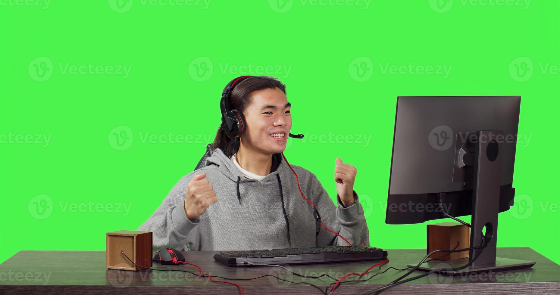 Asian guy sitting at office desktop, having fun with friends while enjoying internet multiplayer games with headphones. Over full body greenscreen, man competes in rpg contest. photo