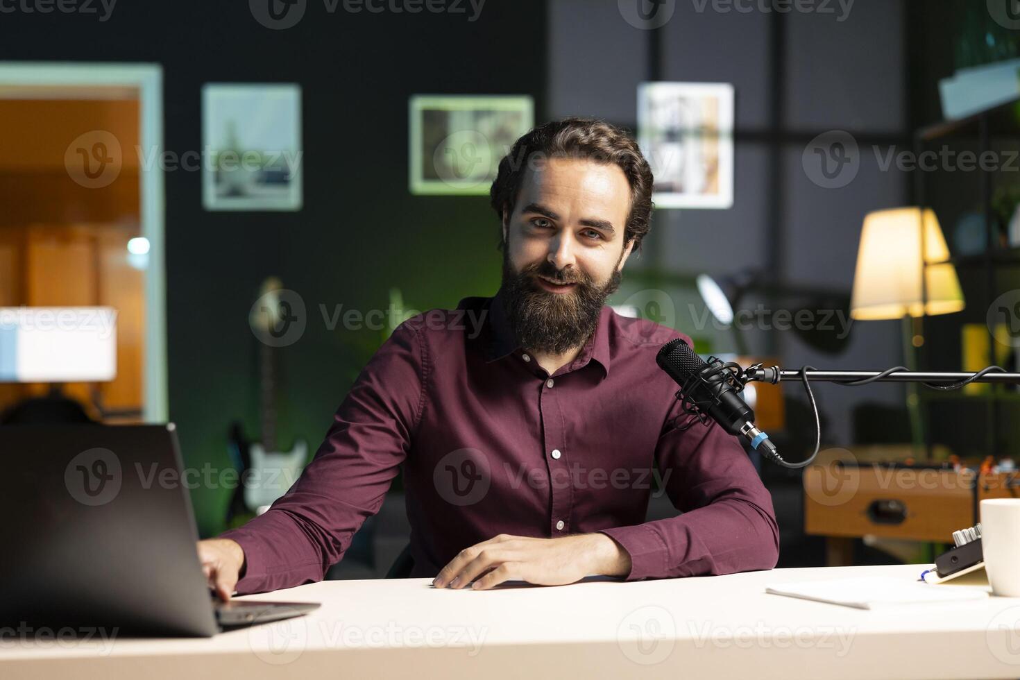 Portrait of smiling man in studio looking at videos on laptop sent by viewers, filming with professional camera. Cheerful entertainer reacting to clips sent by audience during live broadcast photo