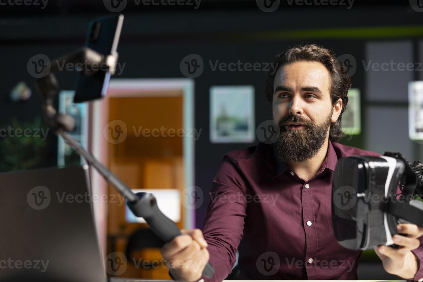 Tech vlogger reviewing VR glasses in apartment studio, recording with phone. Man filming virtual reality headset review vlog for technology enthusiasts using smartphone on selfie stick photo