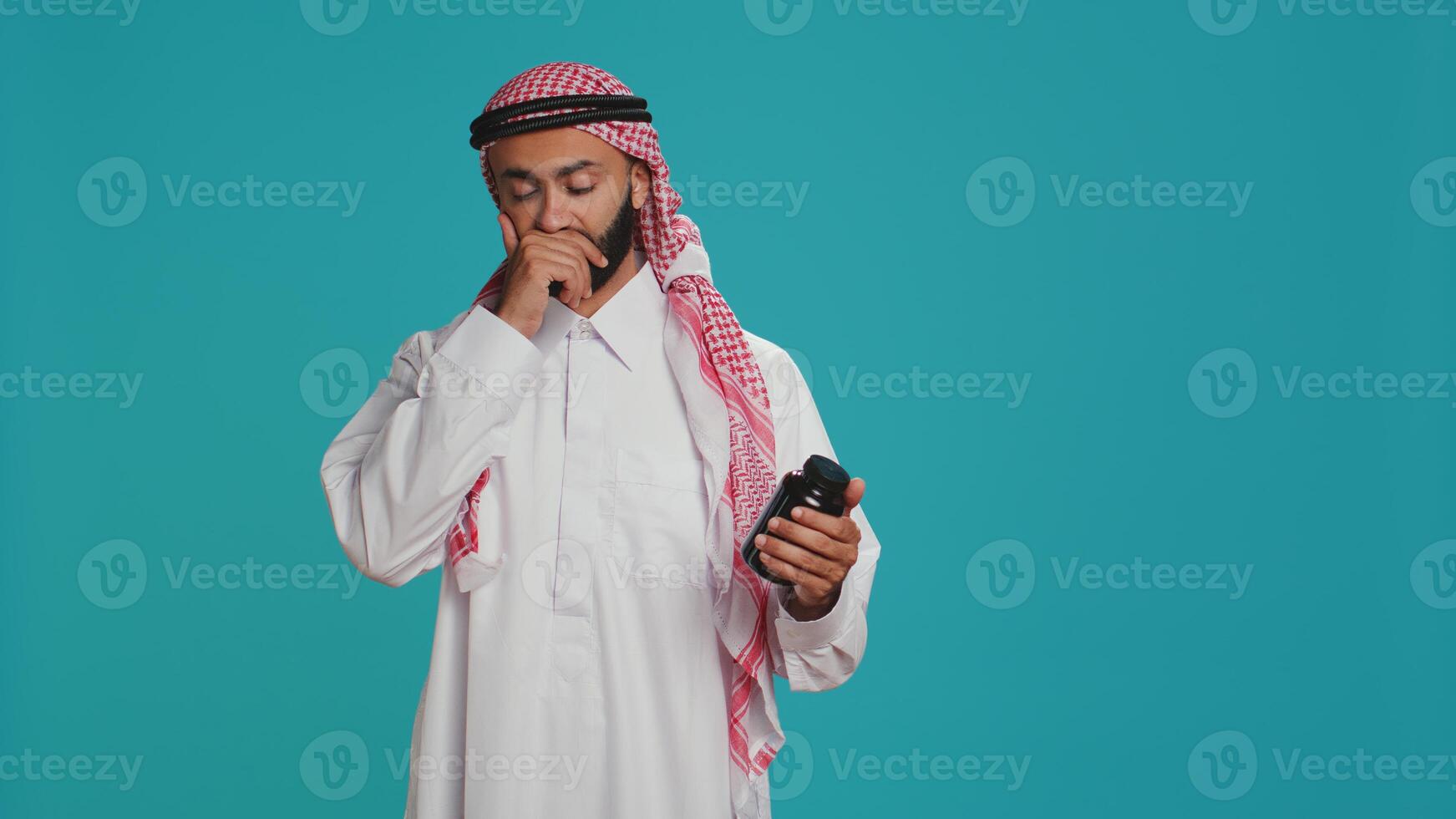 Muslim adult looks at jar of pills on camera, preparing to take painkillers or vitamins from bottle of medicine. Young person examining healthcare drugs in studio, medical treatment. photo