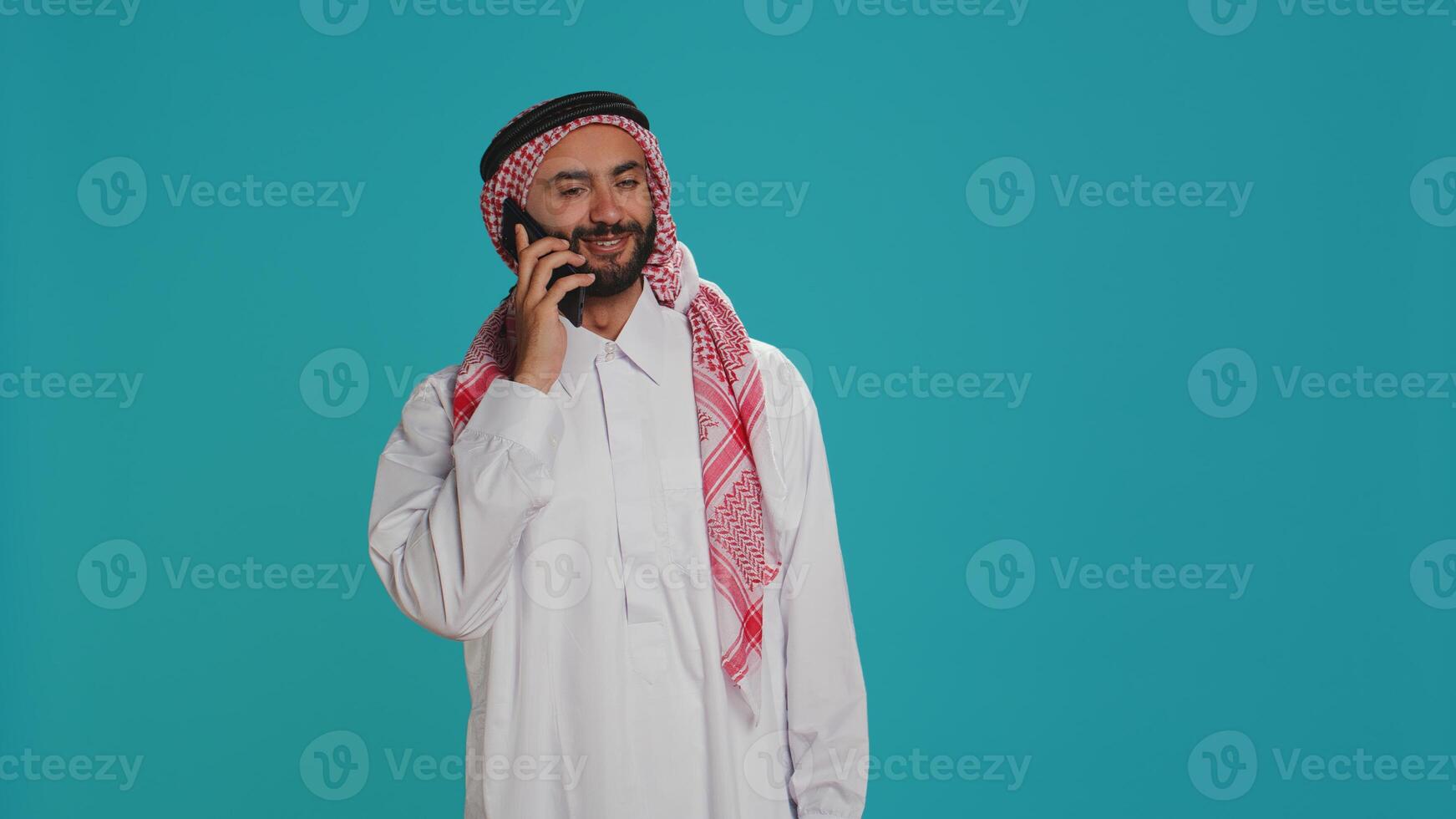 Islamic guy in robe texting on phone while posing with confidence over blue background. Middle eastern person dressed in thobe and headscarf, sending online messages on smartphone app. photo