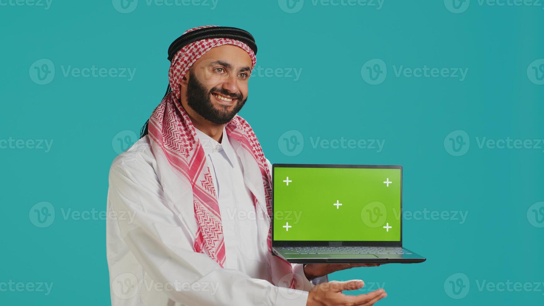 Islamic person shows greenscreen laptop, presenting modern isolated layout for copyspace. Young adult showcasing blank chromakey mockup on wireless pc, wearing thobe and headscarf. photo