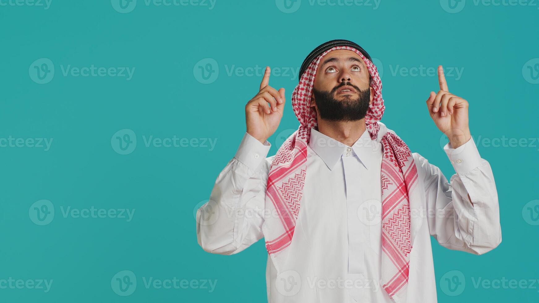 Middle eastern guy looking up above head, pointing and indicating direction upwards in studio. Young islamic person wearing traditional arabic clothing with robe and headscarf, top indication. photo